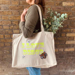 side view of woman carrying cream cotton and polyester tote bag, neon yellow print "I LOVE DISCO" with three iron on patches - a tiger head and 2 silver stars 